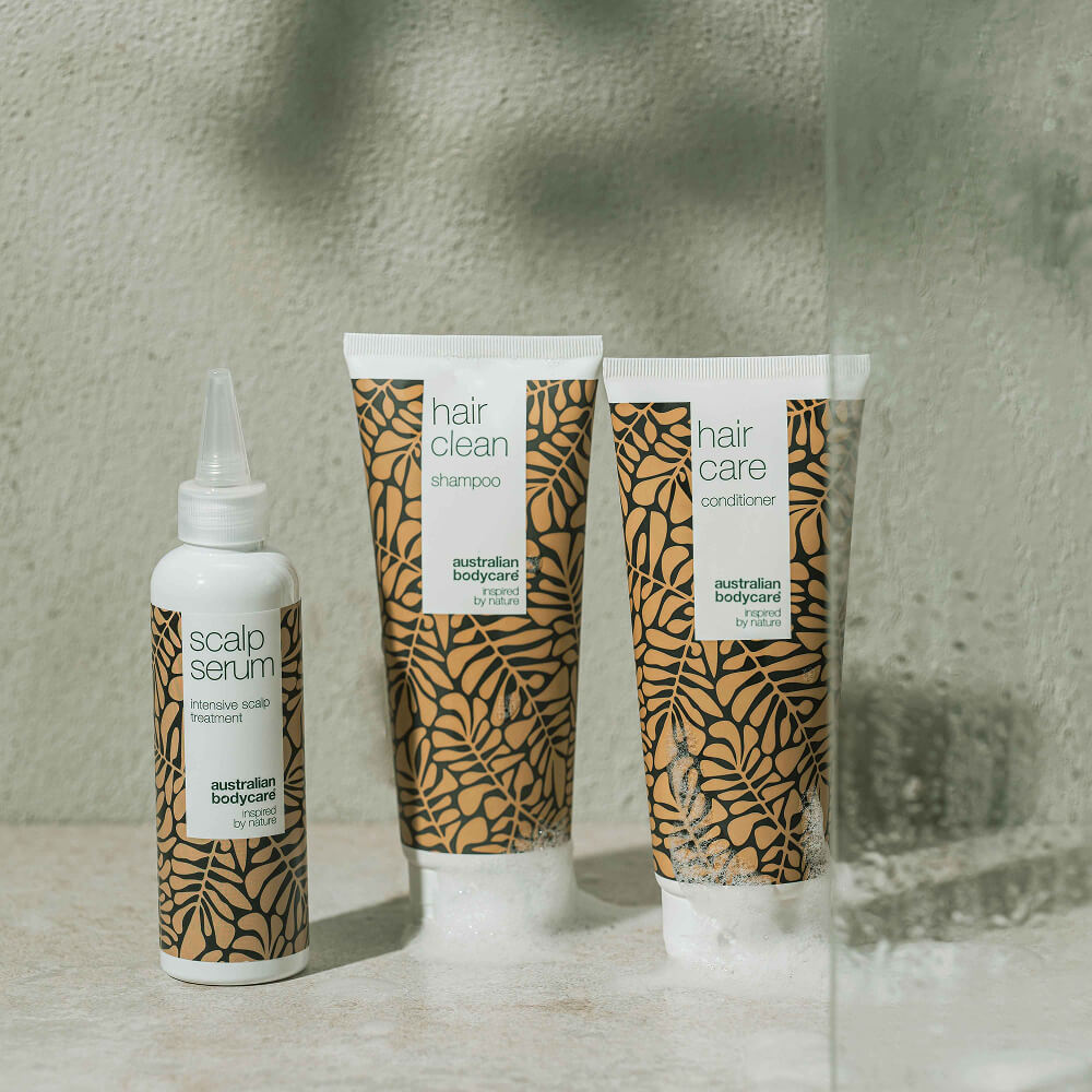 Trois soins capillaires Australian Bodycare – Scalp Serum, Hair Clean Shampoo et Hair Care Conditioner – sont posés sur une surface de salle de bain claire, entourés af vand og skum.